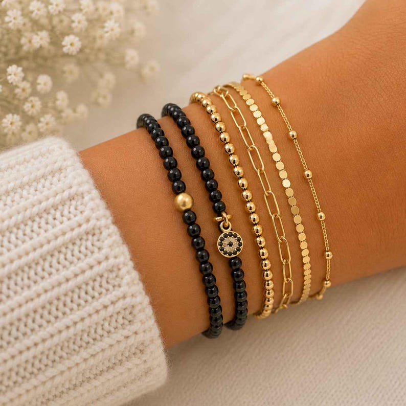 May include: A set of five bracelets featuring two black beaded strands, one with a gold-tone evil eye charm, alongside three gold-tone chains including a beaded strand, a paperclip link chain, and a flat disc chain with small gold spheres.