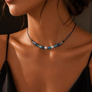 May include: A choker necklace featuring a thin dark blue cord strung with a series of small, round beads in varying shades of blue, teal, and white, accented by tiny silver-toned metallic spacers positioned between the individual beads.