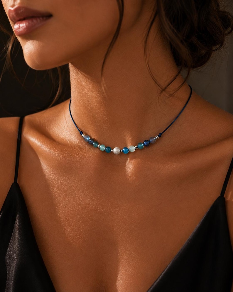 May include: A choker necklace featuring a thin dark blue cord strung with a series of small, round beads in varying shades of blue, teal, and white, accented by tiny silver-toned metallic spacers positioned between the individual beads.