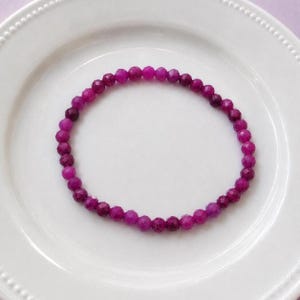 May include: A circular bracelet composed of small, faceted, vibrant magenta-pink beads. The beads are uniform in size and shape, creating a continuous loop. The bracelet is displayed on a plain white surface with a subtle beaded edge detail.