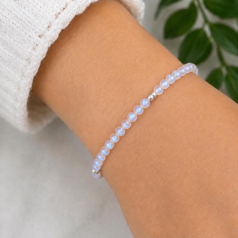 May include: A delicate bracelet composed of small, translucent, iridescent beads with a soft blue glow. The circular beads are strung together in a single strand, punctuated by a single, small, polished silver-toned metal bead near the center.