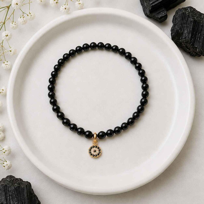 May include: A beaded bracelet featuring smooth, round black onyx beads strung together in a circular pattern. A small, circular gold-toned charm hangs from the center, detailed with a central black stone surrounded by a ring of tiny white crystals.