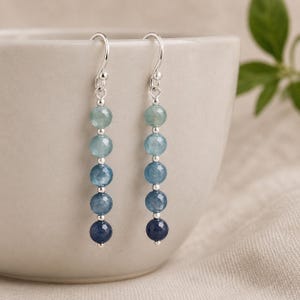 May include: A pair of drop earrings featuring five faceted gemstone beads in an ombre color gradient, transitioning from light aqua to deep navy blue. The beads are separated by small silver-toned metal spacers and hang from silver-toned ear wires.