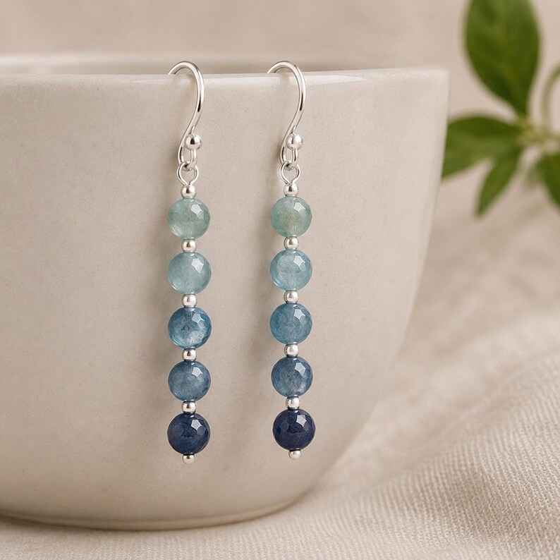 May include: A pair of drop earrings featuring five faceted gemstone beads in an ombre color gradient, transitioning from light aqua to deep navy blue. The beads are separated by small silver-toned metal spacers and hang from silver-toned ear wires.