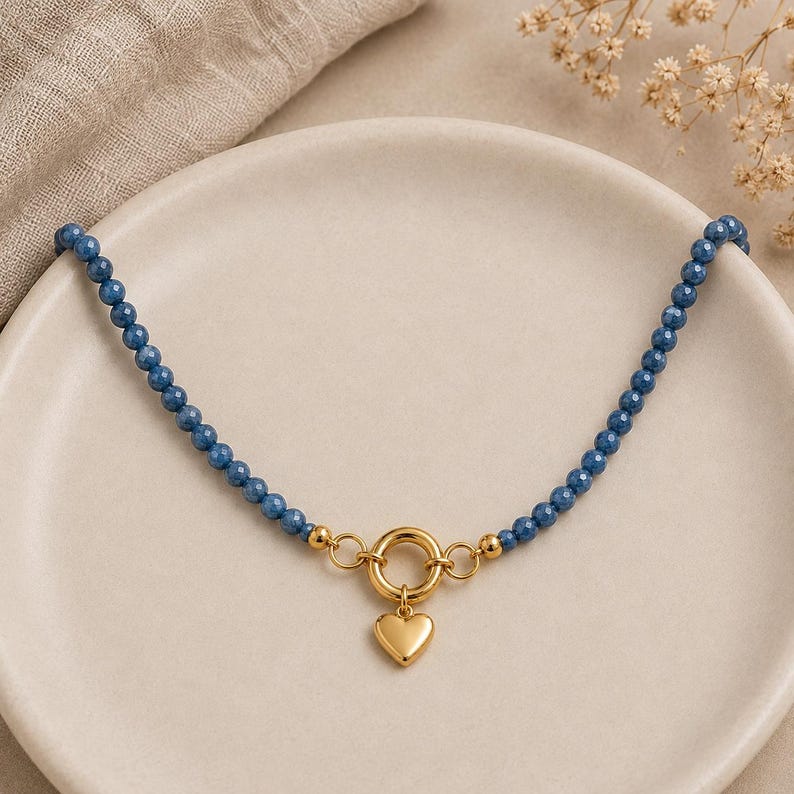 May include: A beaded necklace featuring small, faceted blue gemstone beads arranged in a single strand. The necklace includes a gold-toned circular toggle clasp at the centre, from which a smooth, polished gold-toned heart-shaped charm hangs.