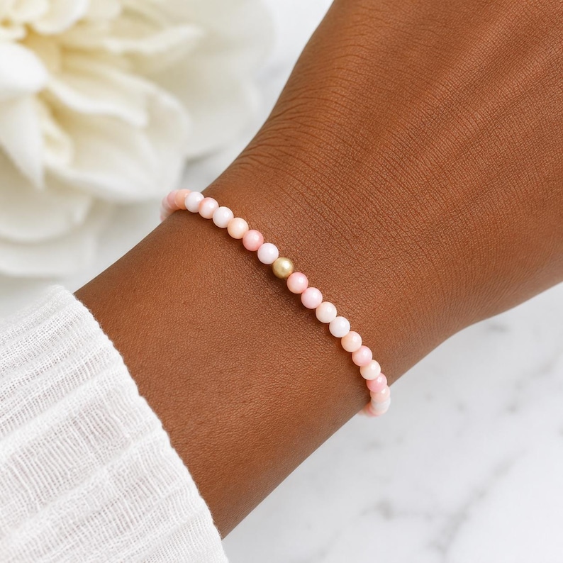 May include: A delicate beaded bracelet featuring small, round, light pink beads arranged in a continuous strand, accented by a single, centred, smooth gold-toned bead. The bracelet is worn on a wrist, showcasing the uniform size of the pink beads.