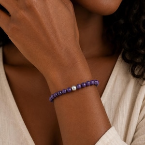 May include: A beaded bracelet featuring small, faceted deep purple stones arranged in a continuous circular band, accented by a single, smooth, polished silver-toned spherical bead positioned prominently among the purple stones.