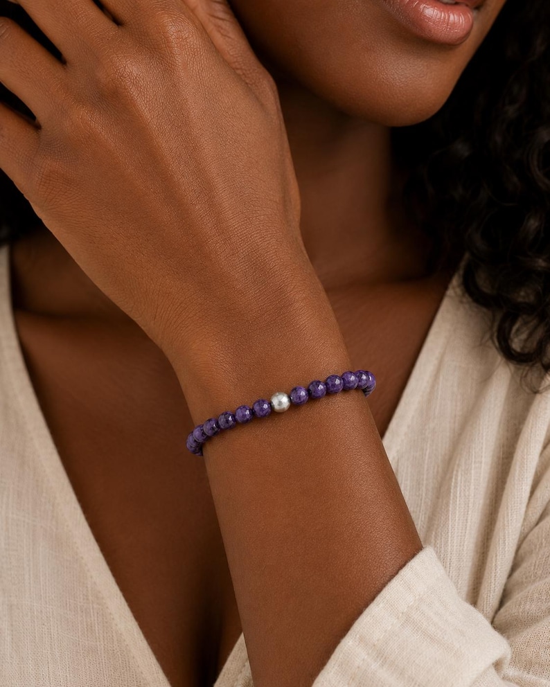 May include: A beaded bracelet featuring small, faceted deep purple stones arranged in a continuous circular band, accented by a single, smooth, polished silver-toned spherical bead positioned prominently among the purple stones.
