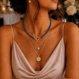 May include: A layered necklace set featuring a strand of small blue beads with a gold circular link, a delicate gold chain with a single round crystal pendant, and a gold chain with a textured circular medallion pendant, plus matching earrings.