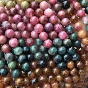 May include: A collection of round, polished tourmaline beads in a variety of colors including shades of pink, deep blue, green, and brown. The beads are strung together on a thin pink cord, showcasing natural variations in tone and translucency.