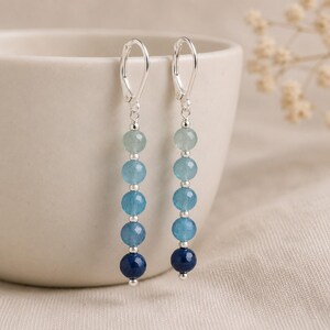 May include: A pair of dangle earrings featuring five faceted round beads in a vertical gradient of blue shades, ranging from light aquamarine to deep navy, separated by small silver-toned metal spacers and finished with silver-toned leverback hooks.