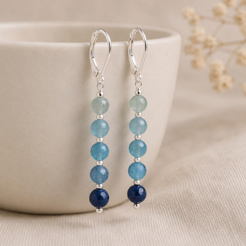 May include: A pair of dangle earrings featuring five faceted round beads in a vertical gradient of blue shades, ranging from light aquamarine to deep navy, separated by small silver-toned metal spacers and finished with silver-toned leverback hooks.