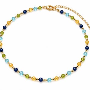 May include: A beaded necklace featuring alternating faceted round beads in shades of dark blue, light blue, yellow, and green, interspersed with small gold-toned metallic spheres. The necklace includes a gold-toned lobster clasp and an extender chain.