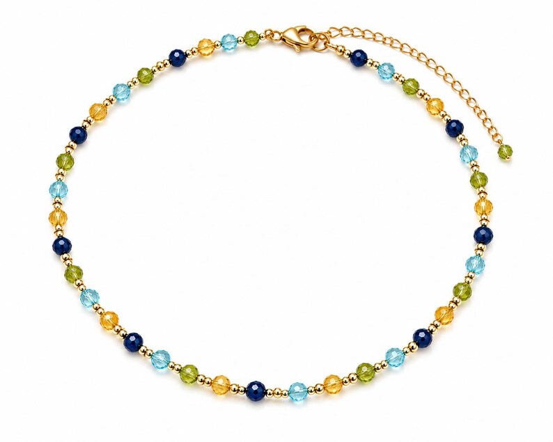 May include: A beaded necklace featuring alternating faceted round beads in shades of dark blue, light blue, yellow, and green, interspersed with small gold-toned metallic spheres. The necklace includes a gold-toned lobster clasp and an extender chain.