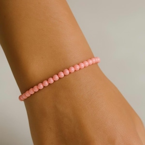 May include: A pink beaded bracelet. The bracelet features small, round beads in a soft, pastel pink hue. The beads are strung together to form the bracelet. The bracelet is worn on a wrist, indicating it is a piece of jewellery.