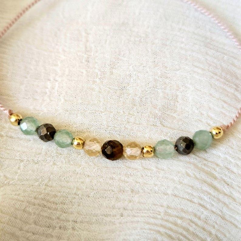 Puede incluir: Una pulsera delicada con un cord&oacute;n fino de color rosa claro ensartado con una secuencia de cuentas facetadas en tonos verde p&aacute;lido, marr&oacute;n oscuro y &aacute;mbar transl&uacute;cido, intercaladas con peque&ntilde;as cuentas esf&eacute;ricas de metal dorado.