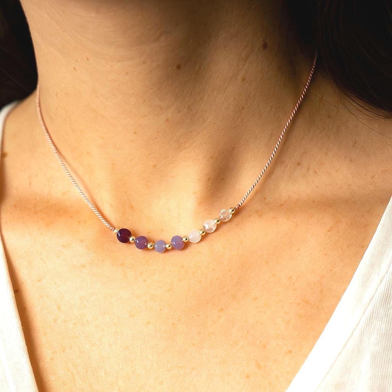 May include: A delicate necklace featuring a thin, light-colored cord and a series of small, round beads in shades of purple, lavender, and white, accented with tiny gold-toned metallic spheres arranged in a row along the center of the strand.