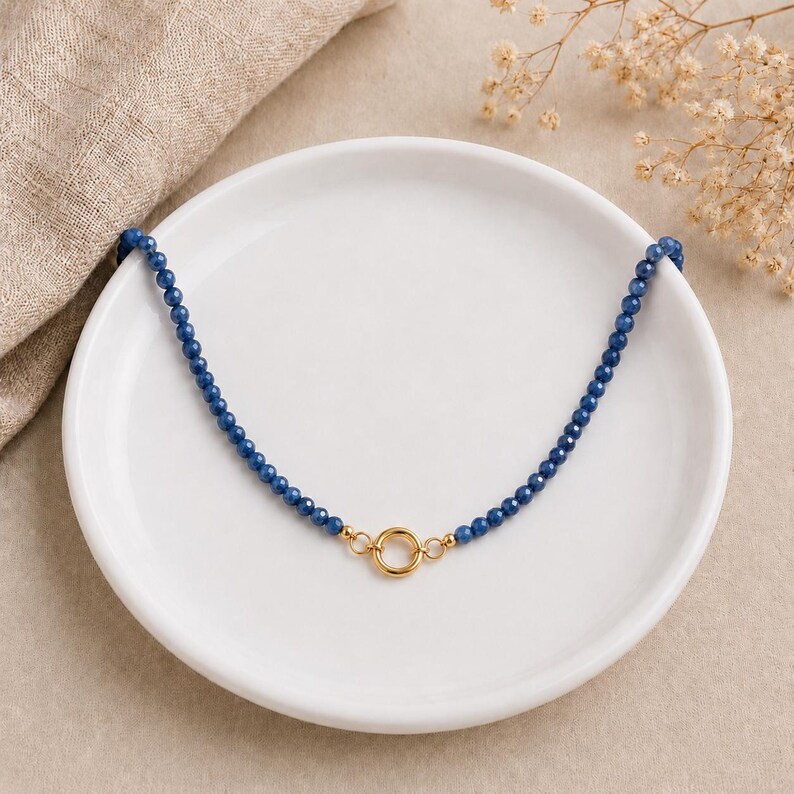 May include: A necklace featuring a single strand of small, faceted, deep blue beads. The two ends of the bead strand are connected by a polished gold-tone circular clasp positioned at the centre, creating a symmetrical and refined jewellery piece.