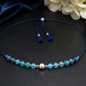 May include: A necklace featuring a dark blue cord with an adjustable sliding closure ending in two small blue tassels. The strand is accented with a central large silver-toned bead, flanked by smaller silver-toned beads and various blue and teal gemstone beads.