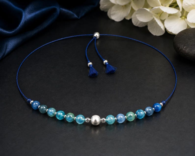 May include: A necklace featuring a dark blue cord with an adjustable sliding closure ending in two small blue tassels. The strand is accented with a central large silver-toned bead, flanked by smaller silver-toned beads and various blue and teal gemstone beads.