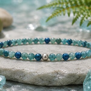 May include: A beaded bracelet featuring small, round, translucent blue and green gemstone beads arranged in a repeating pattern, accented by a single, smooth, polished silver-toned metal bead centered at the front of the circular strand.