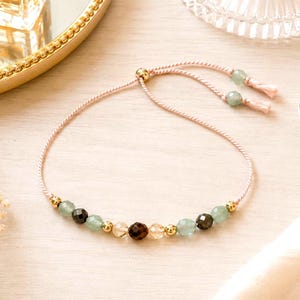 Puede incluir: Una delicada pulsera ajustable con un cord&oacute;n rosa claro y un cierre deslizante. El centro est&aacute; adornado con una secuencia de cuentas facetadas de color verde claro, marr&oacute;n oscuro y transparentes, intercaladas con peque&ntilde;as esferas de metal dorado.