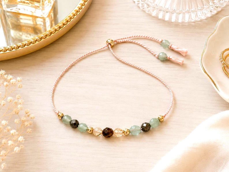 Puede incluir: Una delicada pulsera ajustable con un cord&oacute;n rosa claro y un cierre deslizante. El centro est&aacute; adornado con una secuencia de cuentas facetadas de color verde claro, marr&oacute;n oscuro y transparentes, intercaladas con peque&ntilde;as esferas de metal dorado.