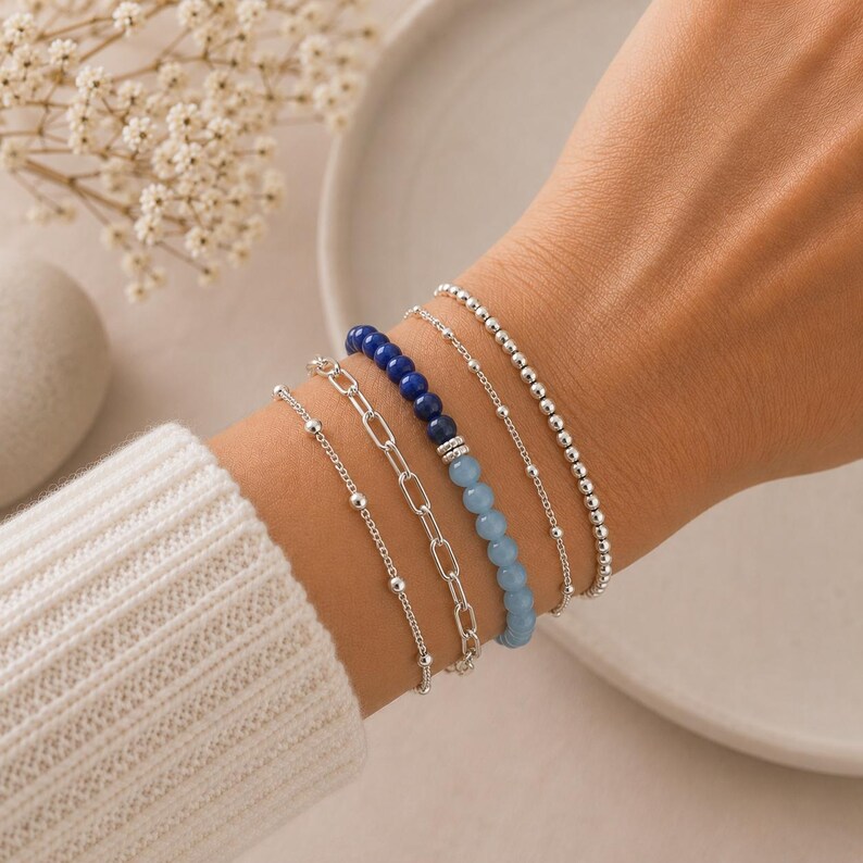 May include: A set of five bracelets worn together. The collection includes a delicate silver bead chain, a silver paperclip link chain, a strand of dark blue and light blue round beads, a thin silver chain, and a silver ball bead bracelet.