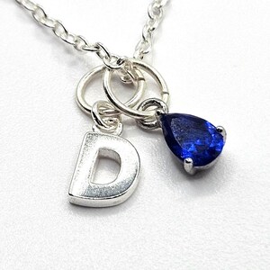 May include: A silver necklace with a silver letter D charm and a teardrop-shaped blue gemstone charm.
