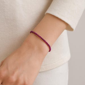 May include: A fine magenta beaded bracelet. The bracelet is made of small, round beads and is worn on a wrist. The background is a soft, off-white colour, providing a neutral backdrop for the jewellery. The bracelet is approximately 17 cm in length.