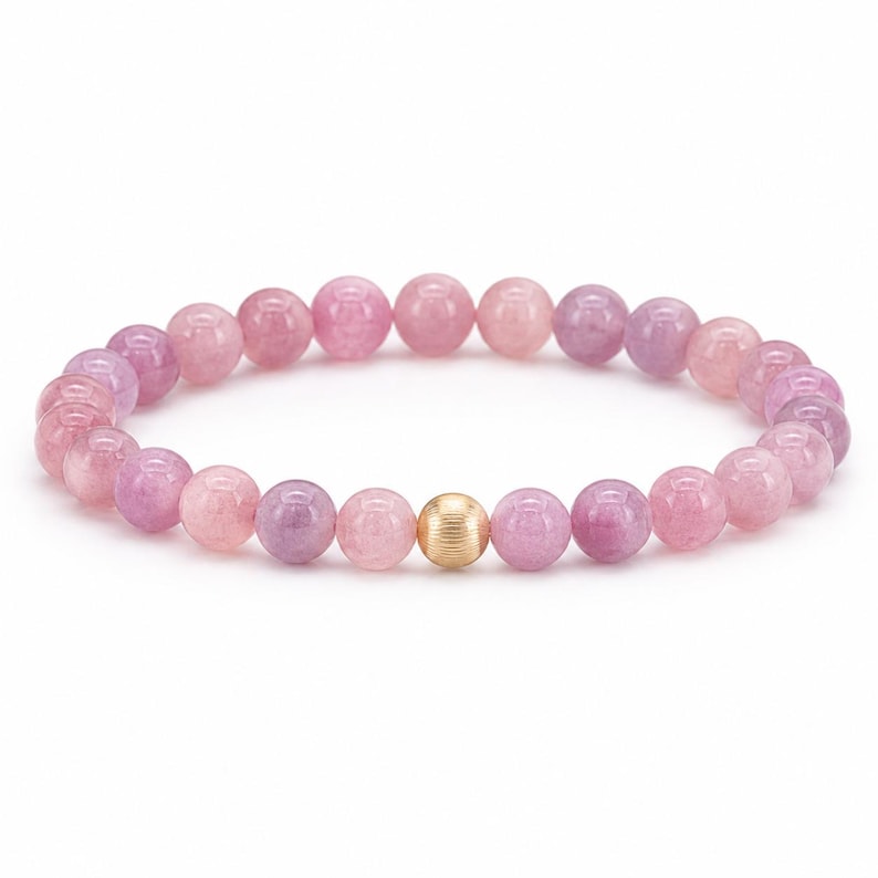 May include: A stretch bracelet composed of smooth, round beads in varying shades of pink and purple. The beads are arranged in a circular pattern, featuring a single, central gold-toned metal bead with a fine, horizontally ribbed texture.