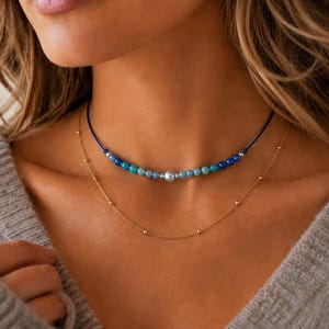 May include: A layered necklace set consisting of a blue cord choker featuring a central silver bead flanked by smaller blue and turquoise beads, and a delicate gold-tone chain necklace with evenly spaced small gold beads along the chain.