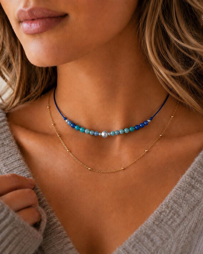 May include: A layered necklace set consisting of a blue cord choker featuring a central silver bead flanked by smaller blue and turquoise beads, and a delicate gold-tone chain necklace with evenly spaced small gold beads along the chain.