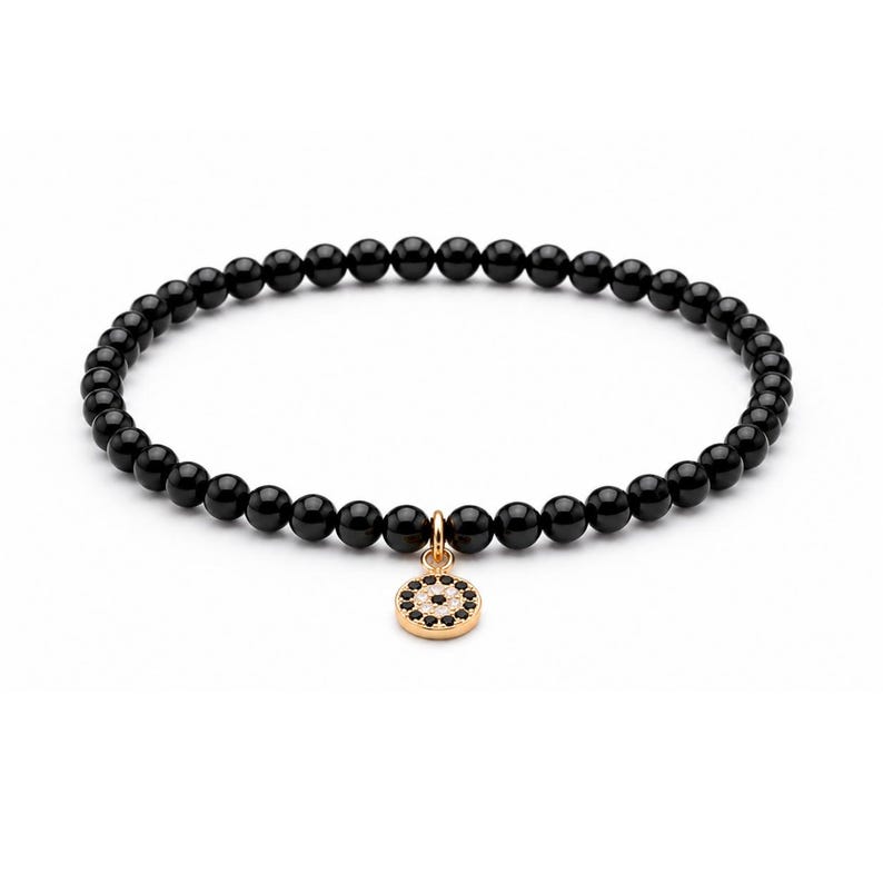 May include: A beaded bracelet featuring smooth, round, polished black beads strung in a continuous circle. A small, circular gold-toned charm hangs from the center, decorated with a pattern of tiny, embedded black and clear white stones.