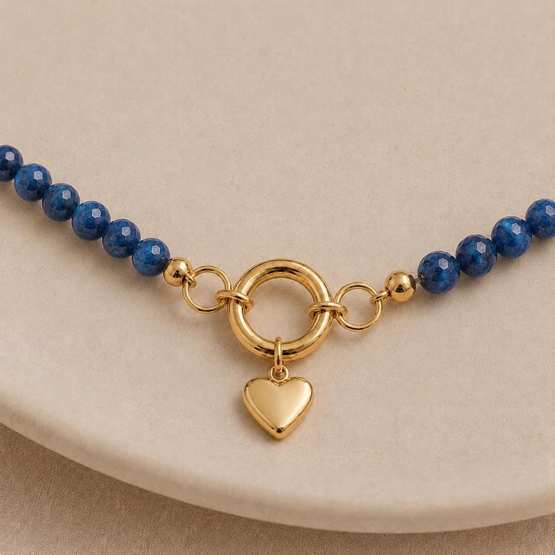 May include: A necklace featuring a strand of faceted deep blue beads connected to a central gold-toned circular clasp. A smooth, polished gold-toned heart-shaped charm hangs from the bottom of the circular ring, creating a focal point.