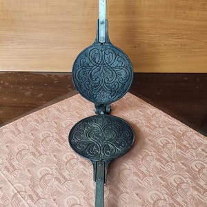 May include: An antique waffle iron with a decorative floral pattern. The iron is made of dark metal and has two round plates connected by a hinge. Each plate features an ornate, symmetrical design. The handles are black.