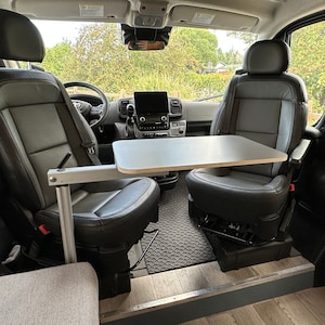 May include: The interior of a van with two black leather seats and a table that folds down from the wall. The table is made of wood and has a light gray finish. The floor is covered in a gray and black patterned mat.