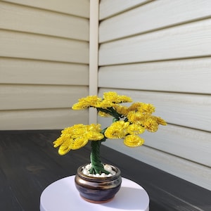 May include: A handcrafted bonsai tree sculpture with bright yellow flowers. The tree has a green twisted trunk and is planted in a small, dark brown ceramic pot filled with white stones. The bonsai is displayed on a white surface.