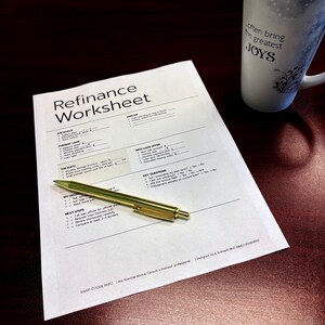 May include: A refinance worksheet with text and a gold pen on a dark wood surface. A white mug with the text "often bring the greatest JOYS" is on the right. The worksheet includes sections for loan details and key questions.