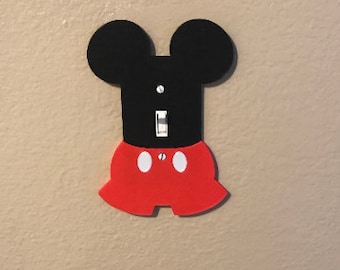 Könnte beinhalten: Ein schwarzer und roter Lichtschalter-Abdeckung mit Mickey Mouse Ohren und Shorts. Die Schalterabdeckung ist so konzipiert, dass sie wie Mickey Mouse aussieht.