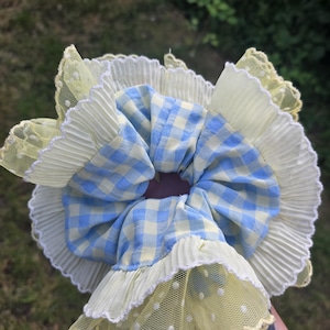 May include: A hair scrunchie featuring a blue and cream gingham fabric centre, surrounded by a ruffled cream pleated trim and an outer layer of sheer pale yellow mesh fabric with a delicate white polka dot pattern and scalloped edges.