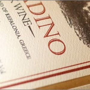 May include: A white wine bottle label with red lettering that reads "Robino Wine" and "Island of Kefalonia, Greece". The label also includes the text "18.2% Vol."