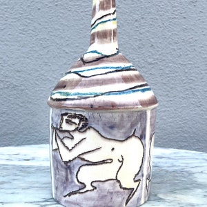 May include: A ceramic vase with a white background and a brown, blue, and white striped design. The vase features a hand-painted image of a nude figure in a reclining pose.