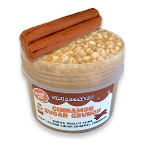 Può includere: Un contenitore in plastica trasparente riempito di slime beige testurizzato con sabbia e perlite, sormontato da un ciondolo marrone a forma di stecca di cannella. L'etichetta riporta: SLIME AFICIONADOS CINNAMON SUGAR CRUNCH, DO NOT EAT.