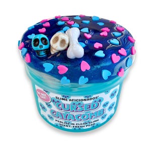 Può includere: Un contenitore in plastica trasparente riempito di slime blu scuro glitterato, decorato con piccoli ciondoli a forma di cuore rosa e azzurri, un teschio blu, un teschio beige e un osso bianco. L'etichetta riporta Slime Aficionados Cursed Catacomb.