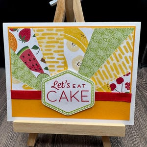 May include: A handmade greeting card with a colorful fruit design and the text "Let's eat cake" in a green circle. The card is on a small wooden easel.