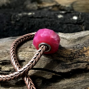 Könnte beinhalten: Eine facettierte, leuchtend pinke Perle mit einem silberfarbenen Kern ist auf einer kupferfarbenen Kette aufgefädelt. Die Perle hat ein geometrisches Design und ist vor einem verwitterten Holzgrund platziert. Die Kette ist im geflochtenen Stil.
