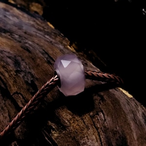 May include: A lilac-coloured, faceted bead with a metallic core is shown on a brown, braided cord. The bead features a geometric design, set against a dark, textured wooden background.
