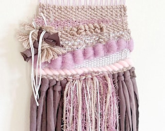 May include: A woven wall hanging with a pink, brown, and white color scheme. The hanging features a variety of textures, including fringe, pom-poms, and woven fabric.