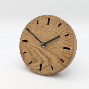 Könnte beinhalten: Eine Holzuhr mit schwarzen Stunden- und Minutenzeigern. Das Zifferblatt der Uhr ist aus Naturholz mit einer Maserung. Die Uhr hat ein schlichtes Design mit schwarzen Stundenmarkierungen.
